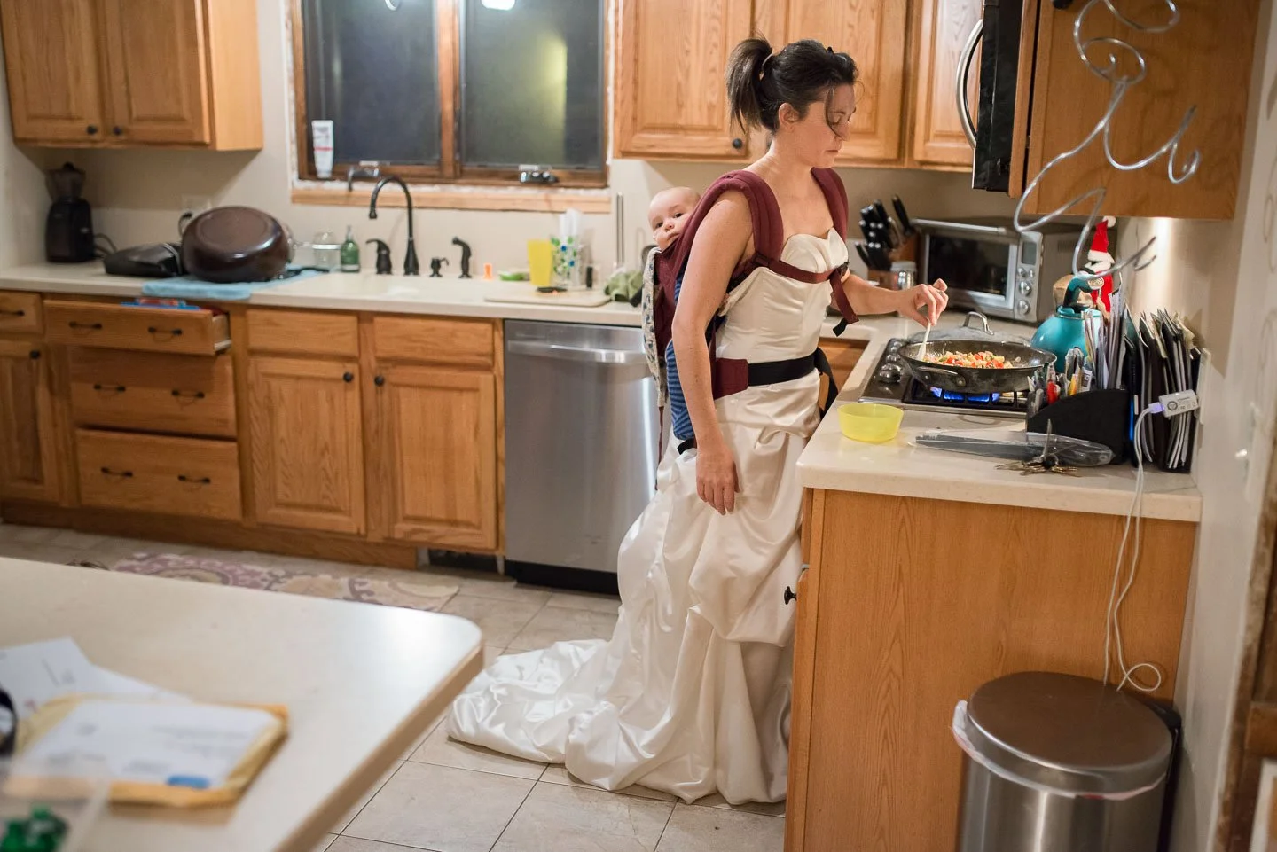 A woman wearing a wedding dress and apron with a baby on her back, cooking in a kitchen.