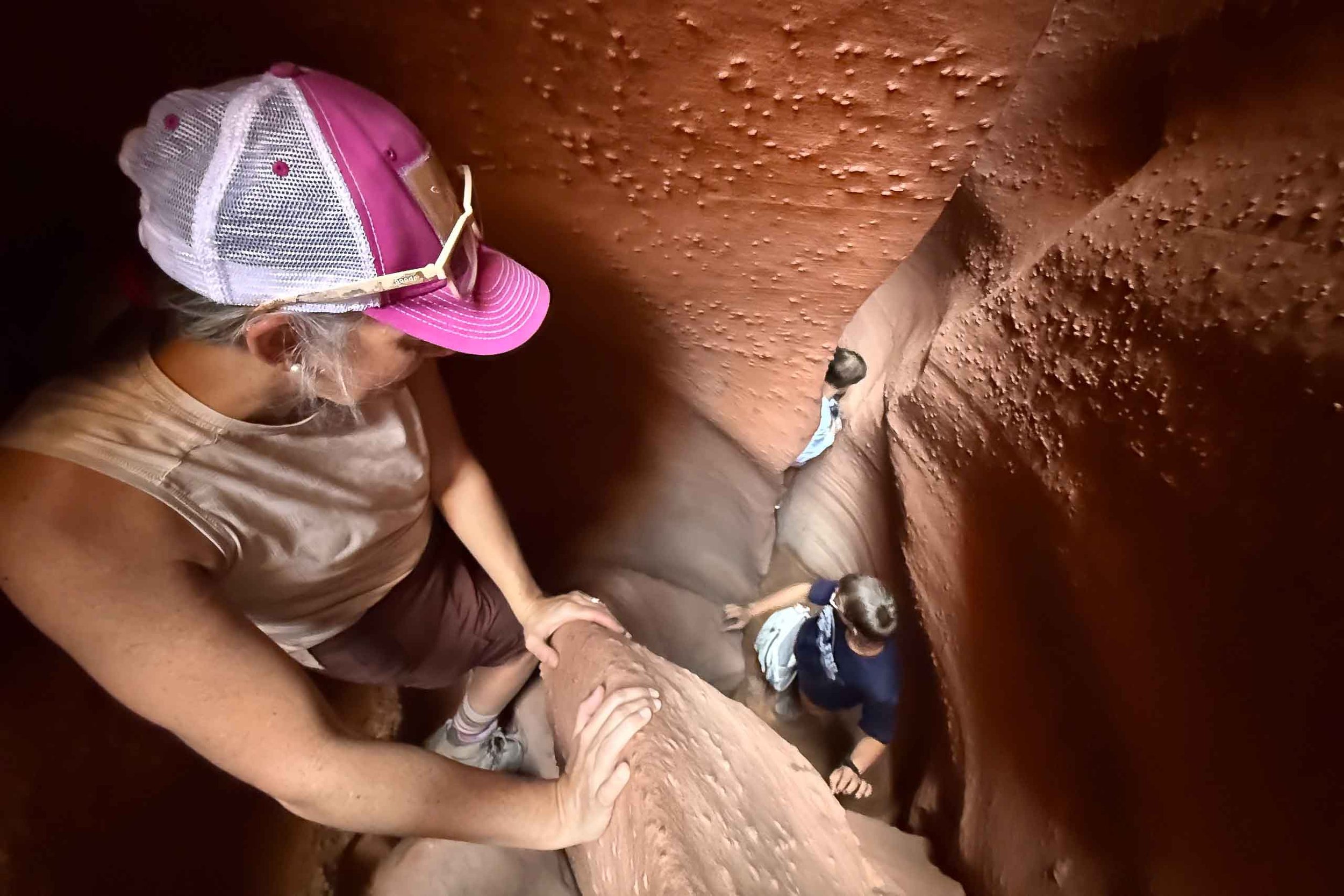 Two children and an adult exploring a slot canyon with red rock walls, climbing down a narrow, winding trail.
