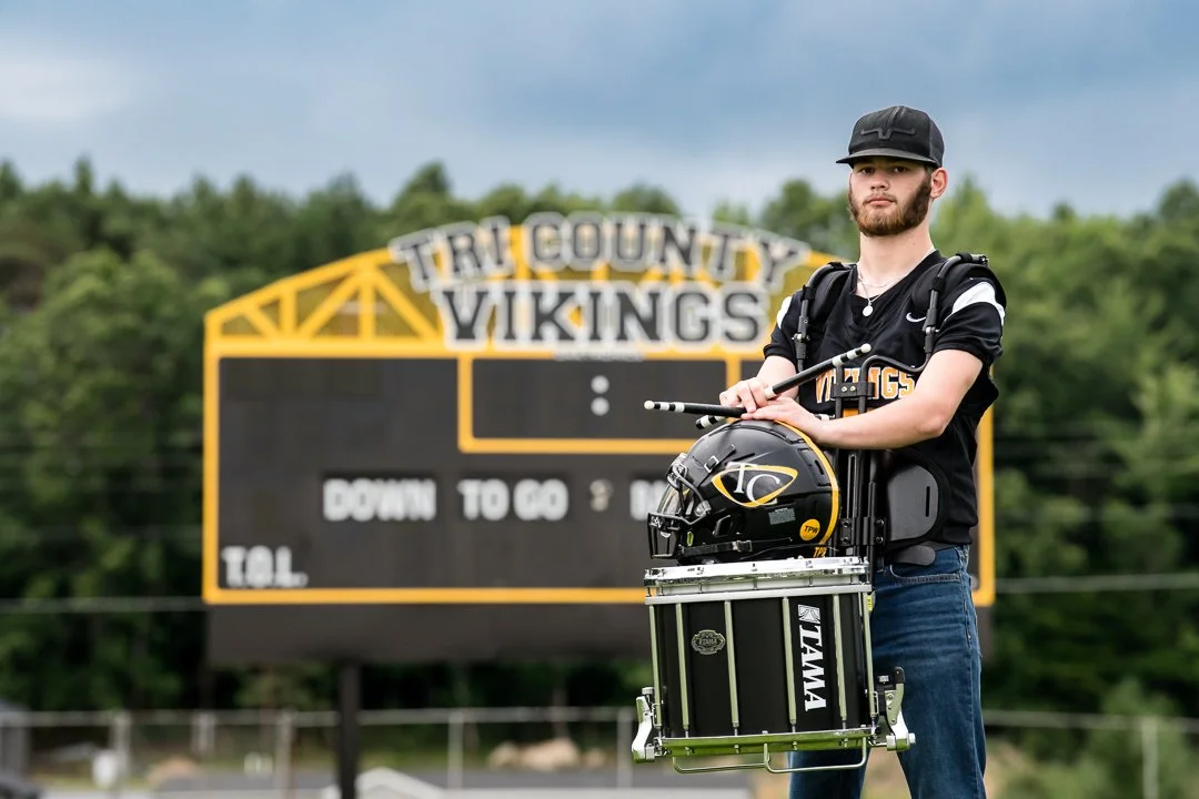 Senior drummer holding a football helmet and snare drum in front of the Tri County Vikings scoreboard