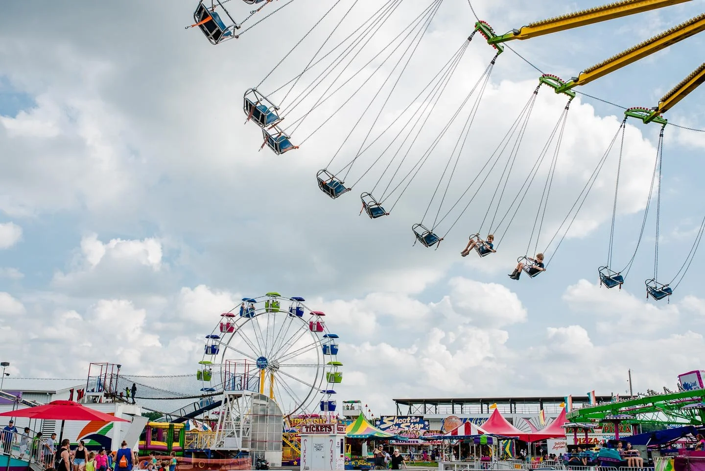 Children riding on swings high in the sky at a carnival with a Ferris wheel and colorful tents and booths below.