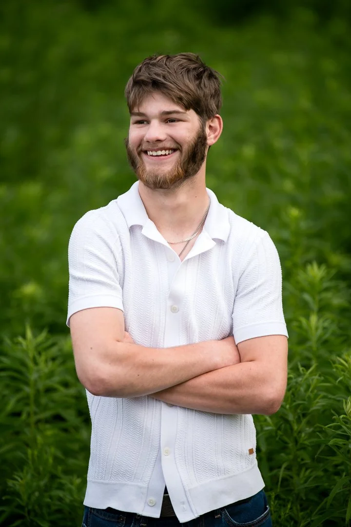A smiling young man with a beard, wearing a white polo shirt, outdoors in front of a green field background — an example of a classic, put-together outfit for a Grand Rapids senior portrait session.