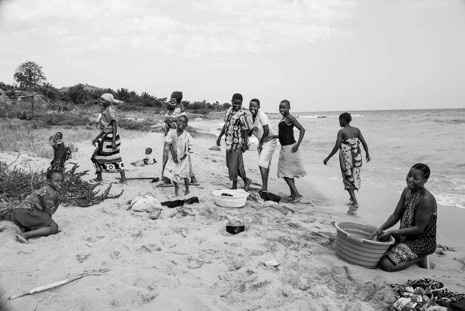 Children and adults at the beach, some standing by the water, others sitting or crouching on the sand, with boats and vegetation in the background.