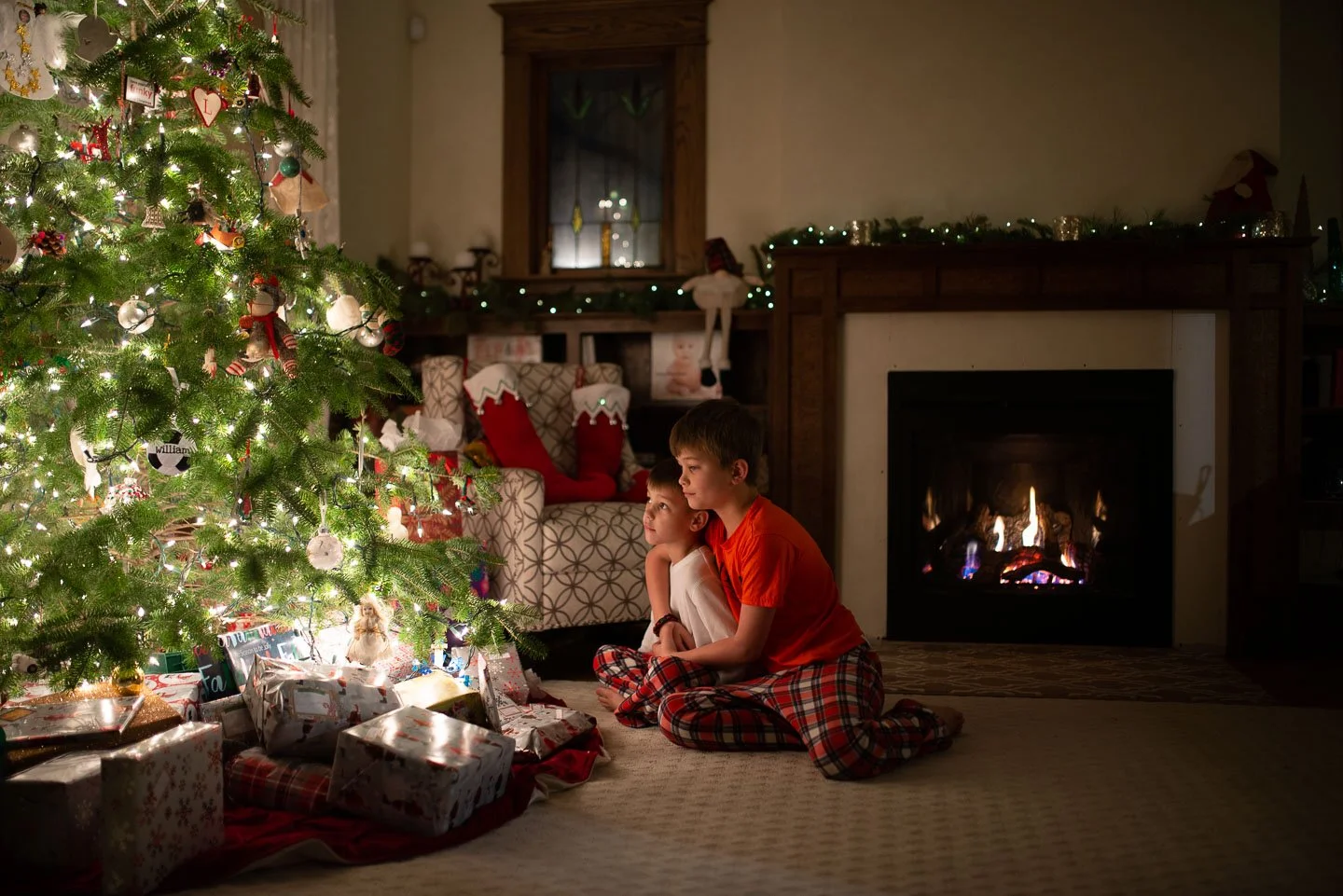 Two children, a boy and a girl, sitting on the carpeted floor in pajamas near a decorated Christmas tree with wrapped presents underneath, in a cozy living room with a lit fireplace and holiday decorations.