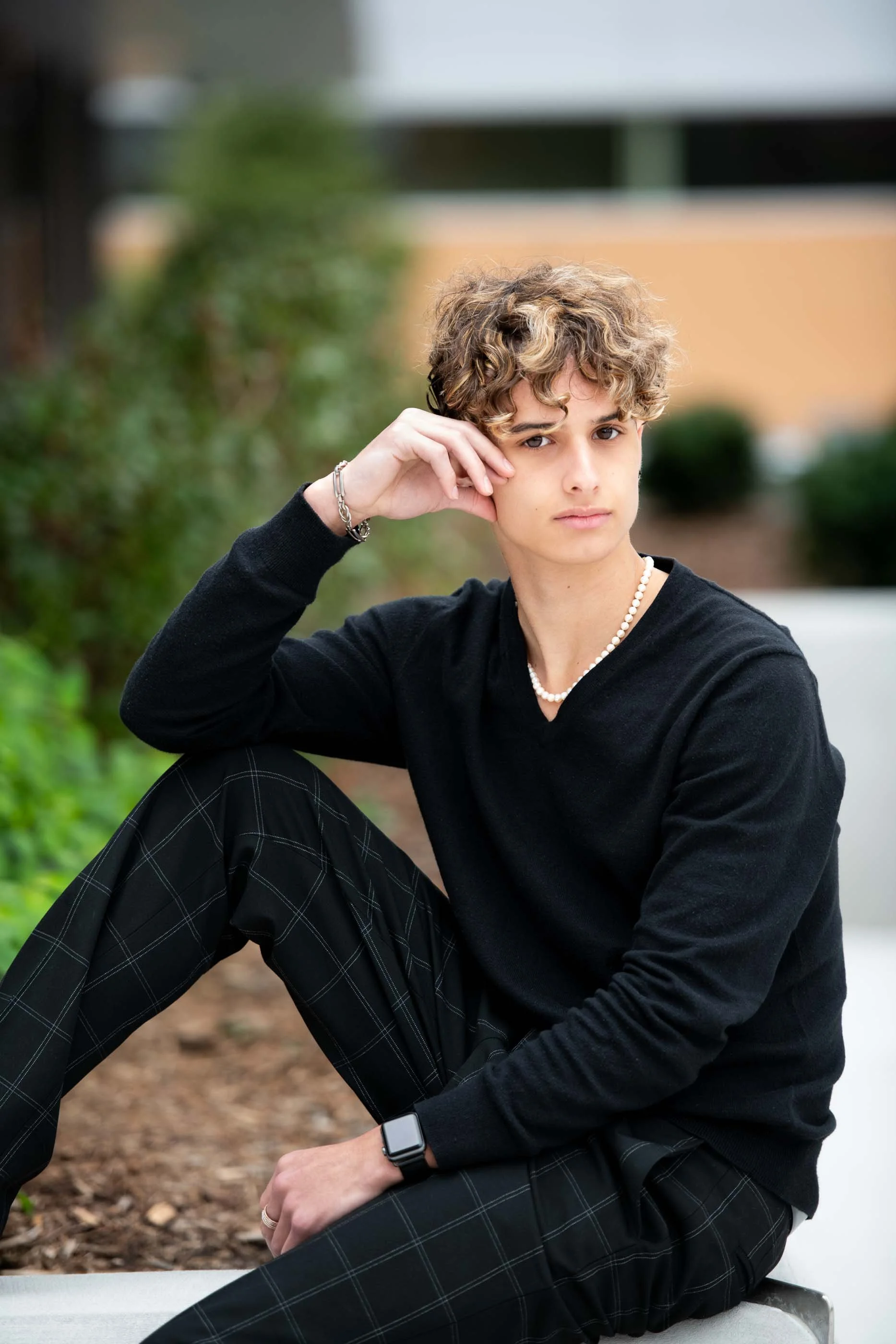 A young person with curly blonde hair sits outdoors on a bench, wearing a black long-sleeve shirt, plaid pants, and accessorized with a pearl necklace, a bracelet, and a smartwatch. The person looks thoughtfully at the camera with one hand resting on their face.