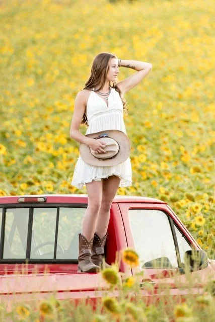 A woman in a white summer dress standing on the hood of a red truck in a sunflower field, holding a hat and looking to the side.