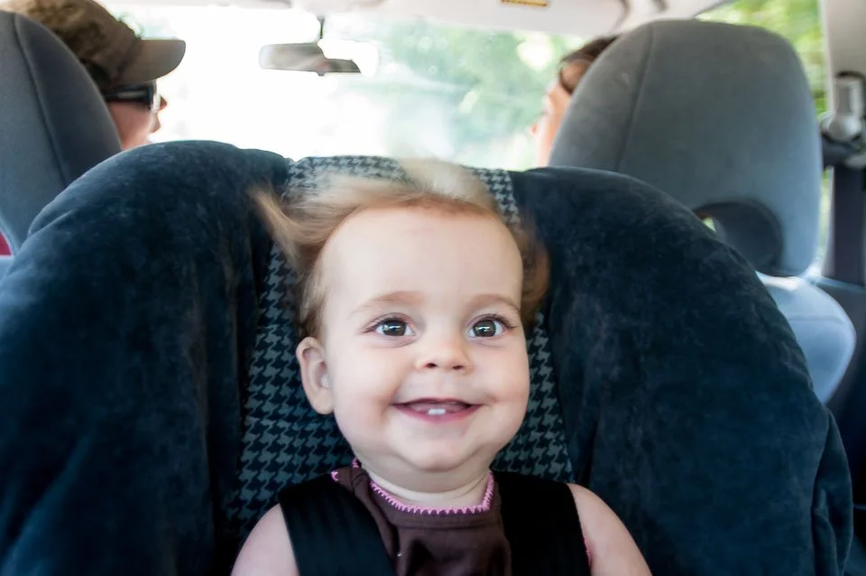 A smiling young girl with light brown hair seated in a car seat in the back of a vehicle, with car seats and other passengers visible in the background.
