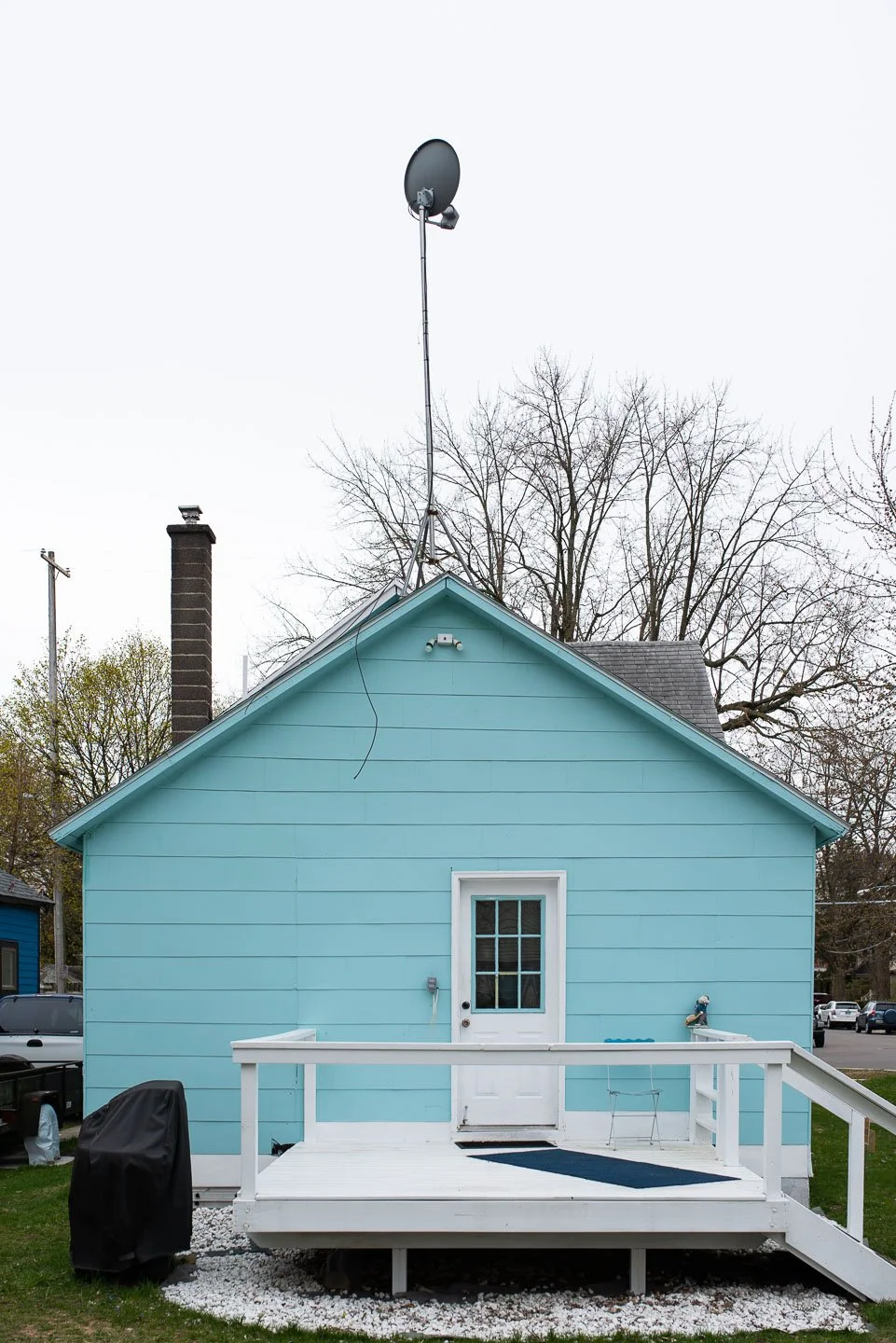 Light blue house with a small deck, a door with a window, and a large satellite dish mounted on the roof, with leafless trees in the background.