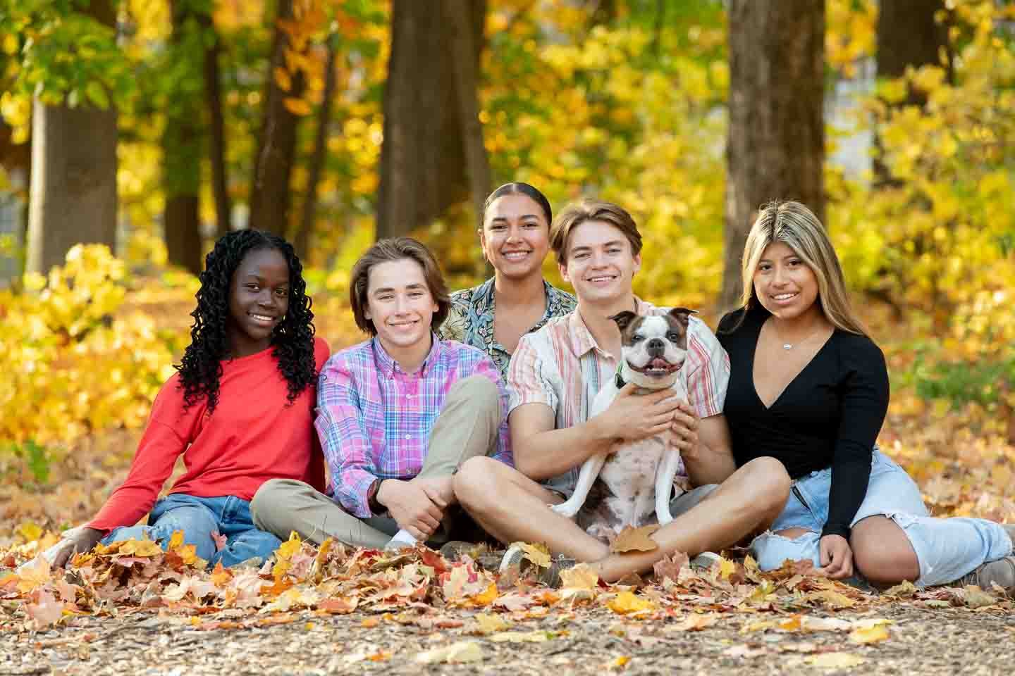 Five young people and a dog sitting on fallen autumn leaves in a forest.