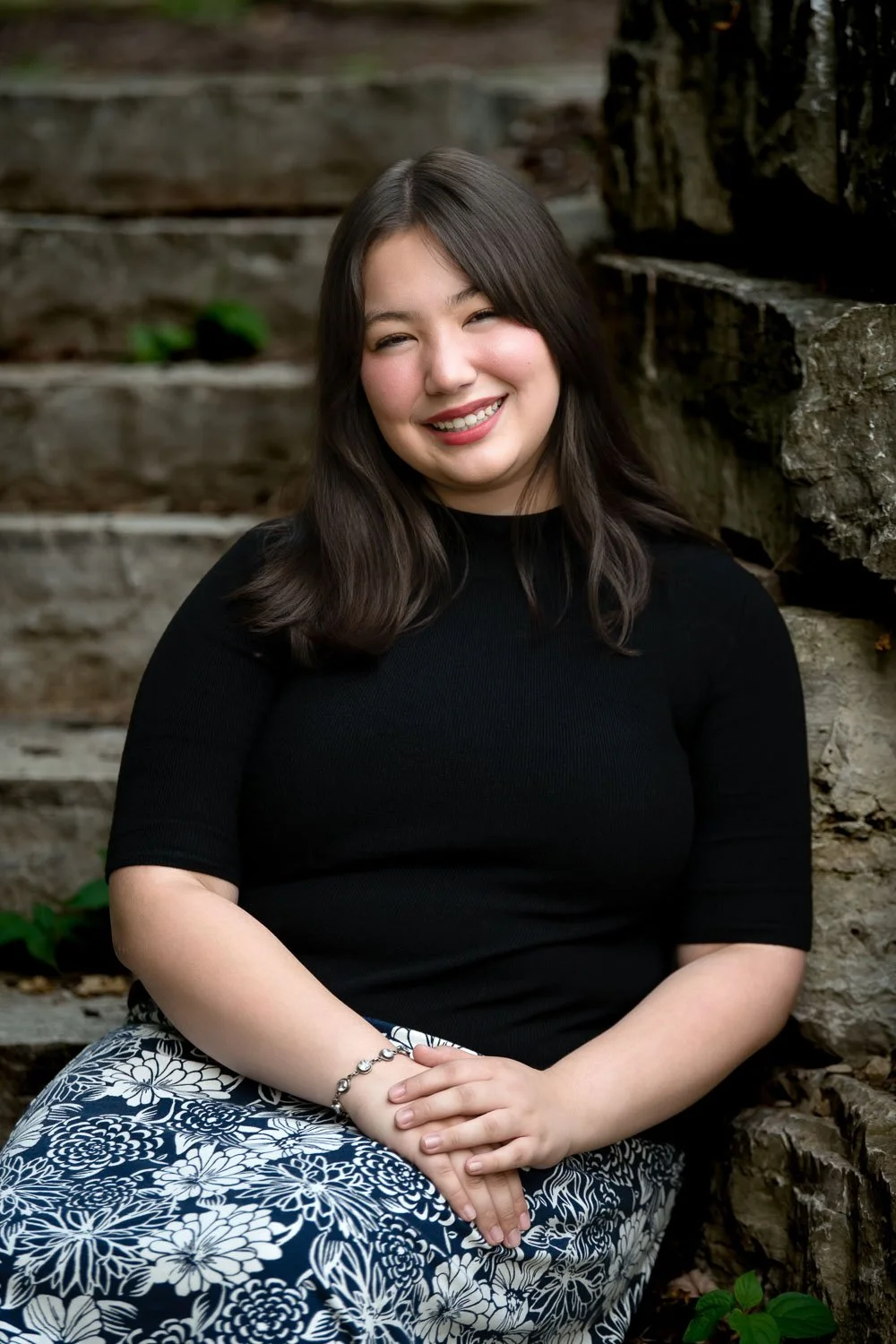 A young woman with shoulder-length dark hair smiling while sitting outdoors against a stone wall with stairs in the background. She is wearing a black top and patterned skirt, with a bracelet on her wrist.