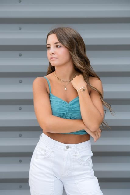 Young woman standing in front of a gray metal wall, wearing a blue crop top and white jeans, with long wavy hair, looking to the side.