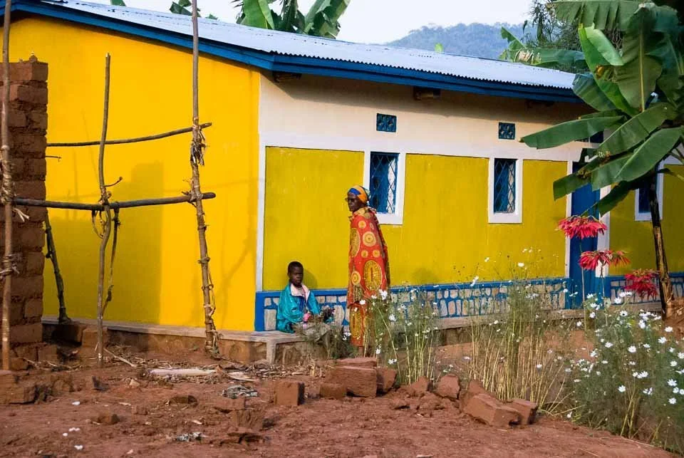 A woman and a child outside a brightly painted yellow house with a blue roof, surrounded by flowers and banana trees.