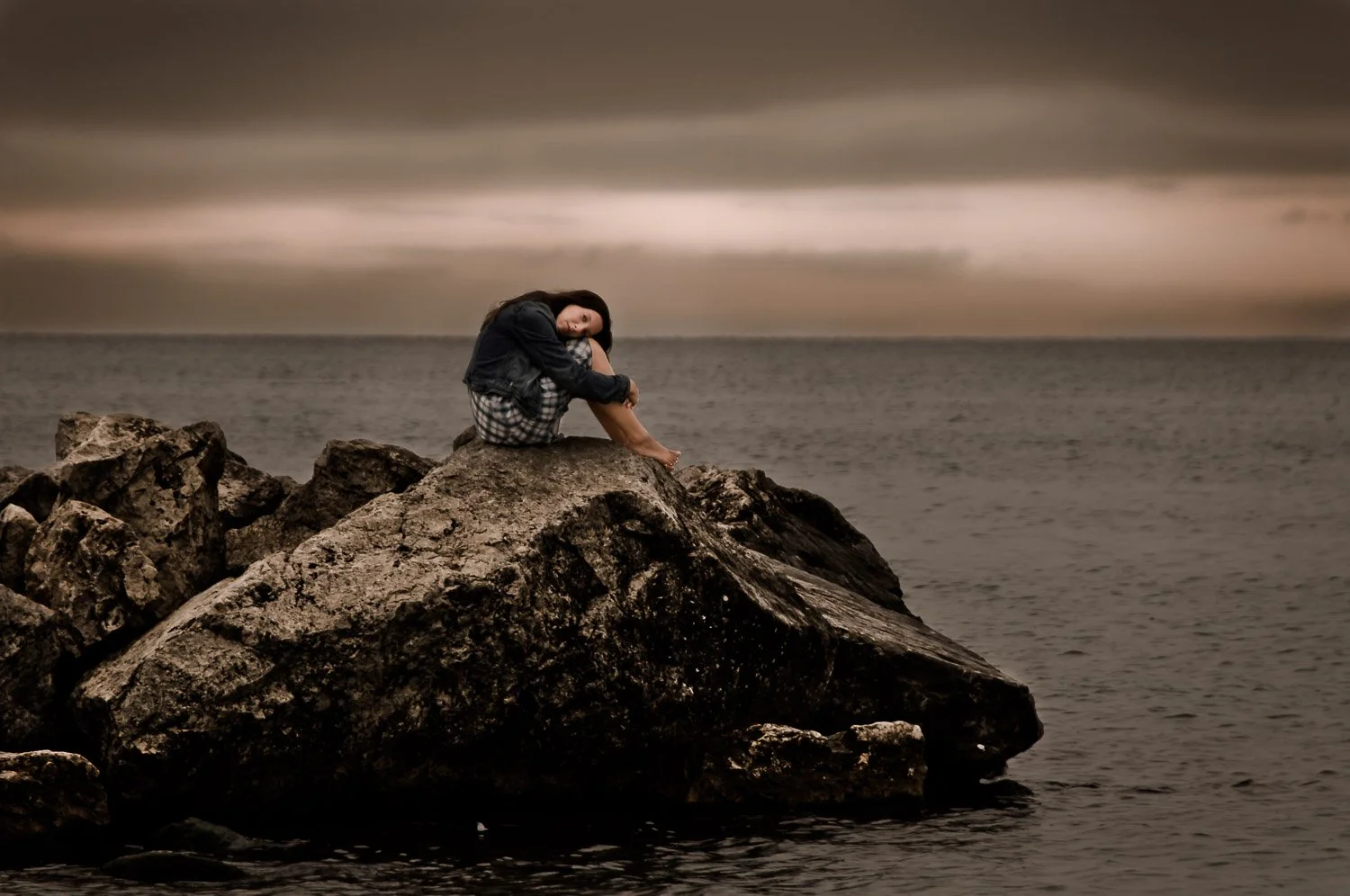 A woman sitting alone on a large rock by the sea at sunset or dusk, with an overcast sky in the background.