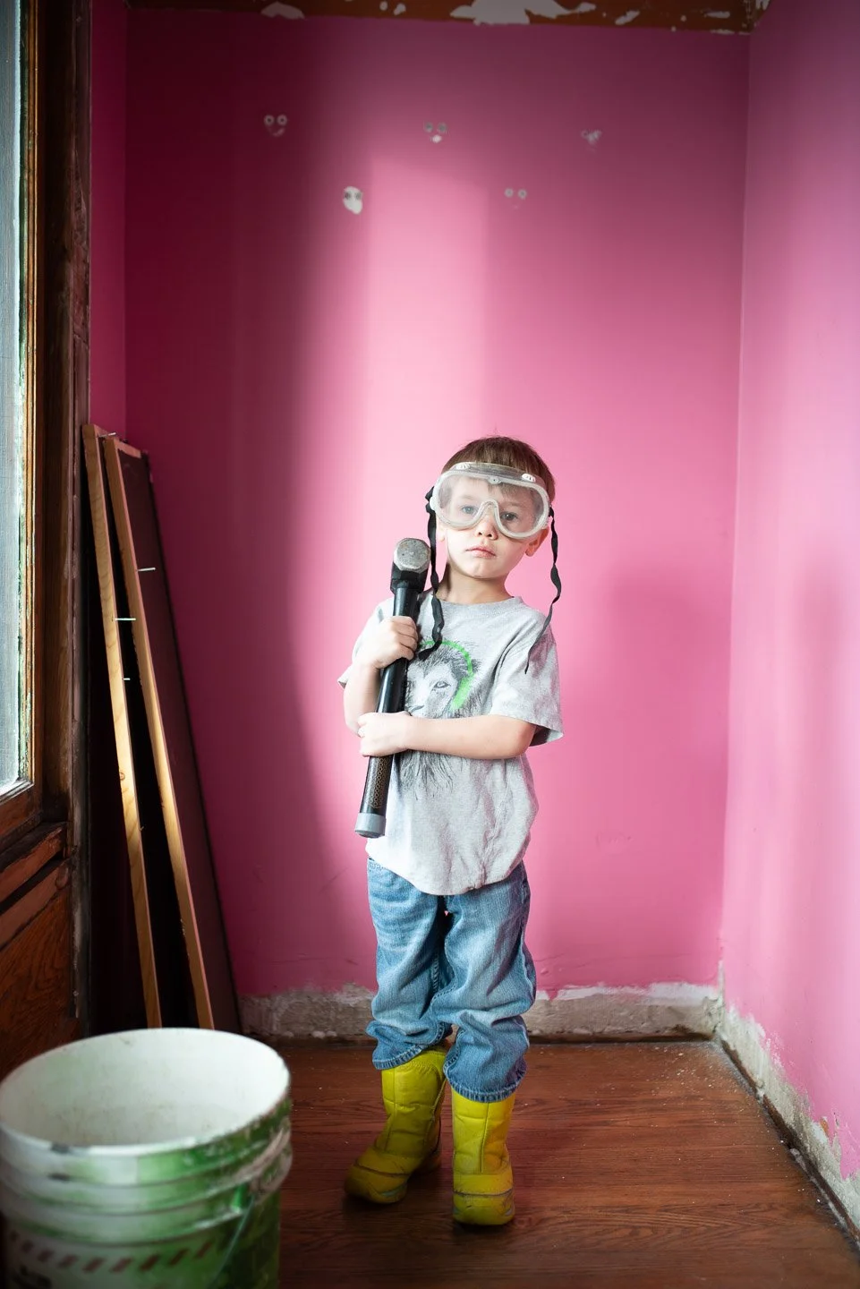 A young boy standing in a room with pink walls, wearing yellow rain boots, blue jeans, a gray t-shirt with a dog graphic, safety goggles, and holding a paint roller, with a bucket of green paint nearby.