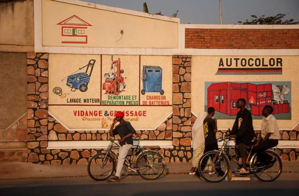 A wall with advertisements for auto repair services, including engine washing, tire repair, and battery charging. There are three people walking and riding bikes in front of the wall.