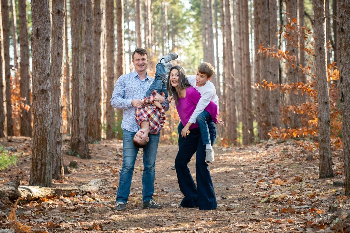 A family of four having fun in a forest, with two parents holding their children upside down and all smiling.