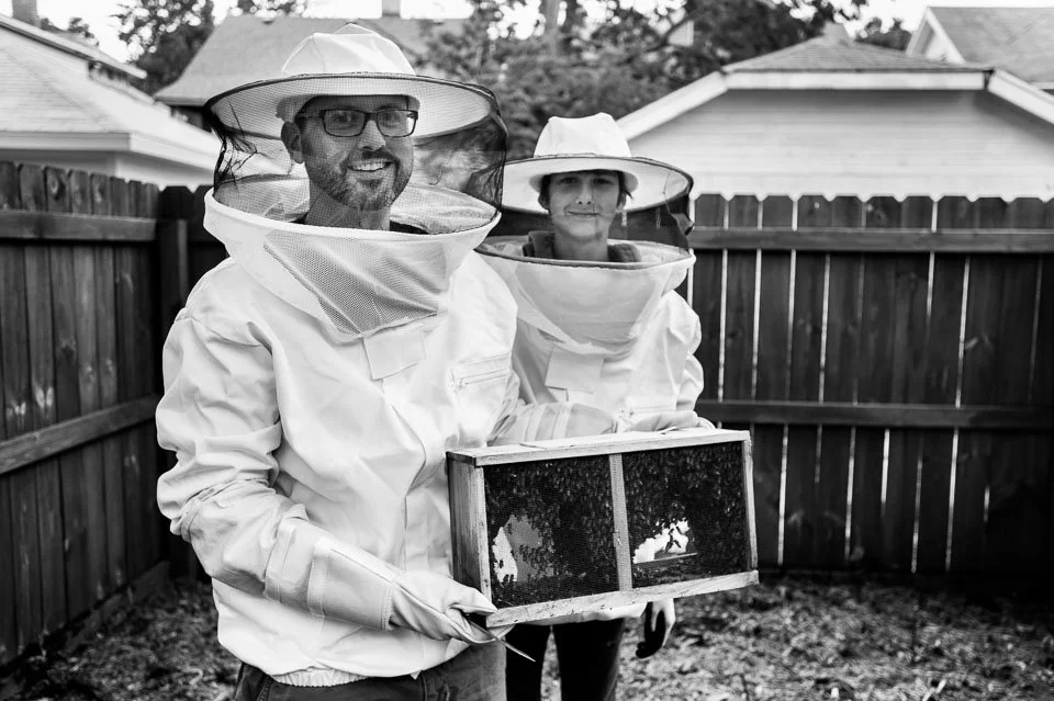 Two people wearing beekeeping suits and hats with veils, holding a honeycomb frame, standing in a backyard with a wooden fence and houses in the background.