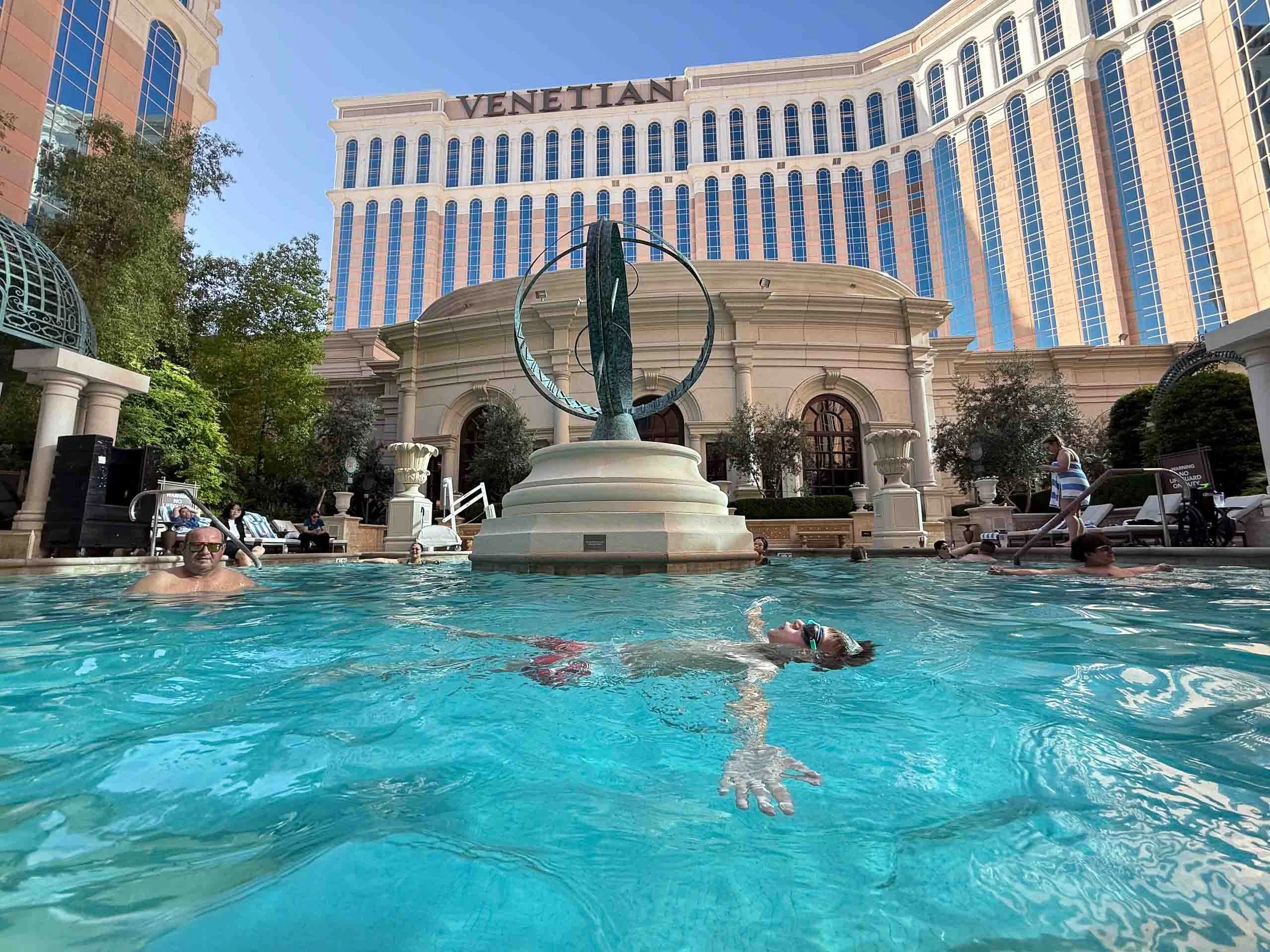Swimming pool scene in front of the Venetian hotel, with people swimming and lounging, surrounded by trees and classical architecture, under a clear blue sky.