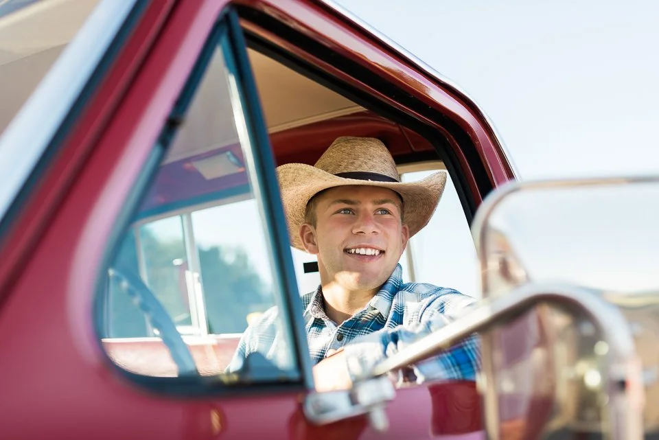 A smiling young man wearing a cowboy hat and a plaid shirt sitting inside a classic red pickup truck.
