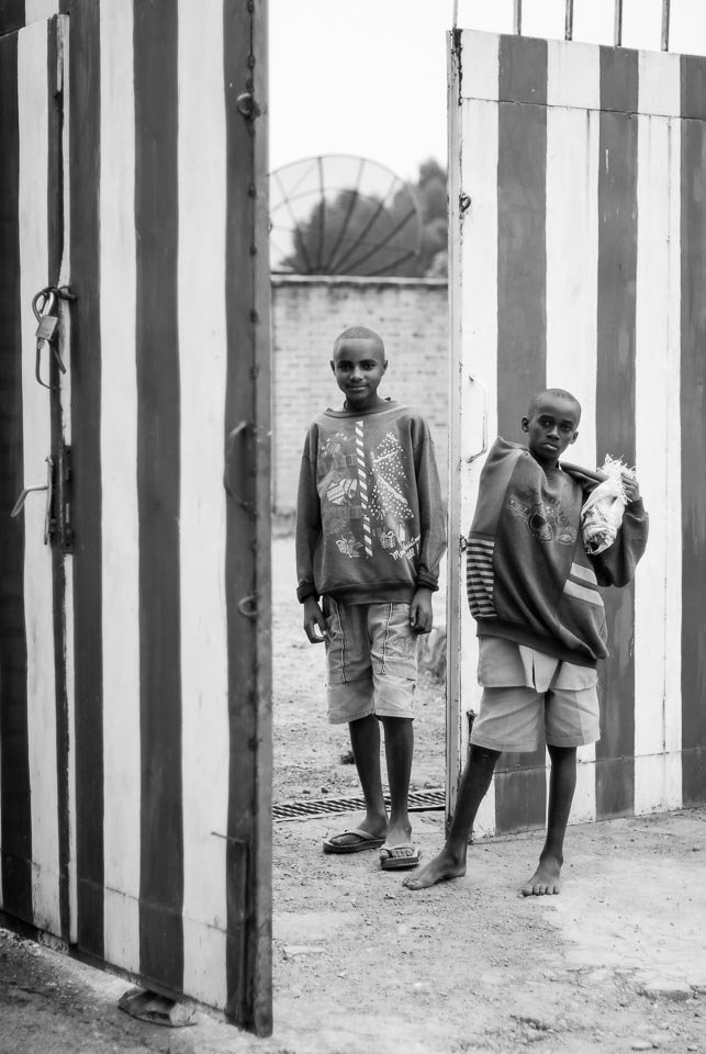Two young boys standing outside a large striped gate, one holding a plastic bag, in a residential area.