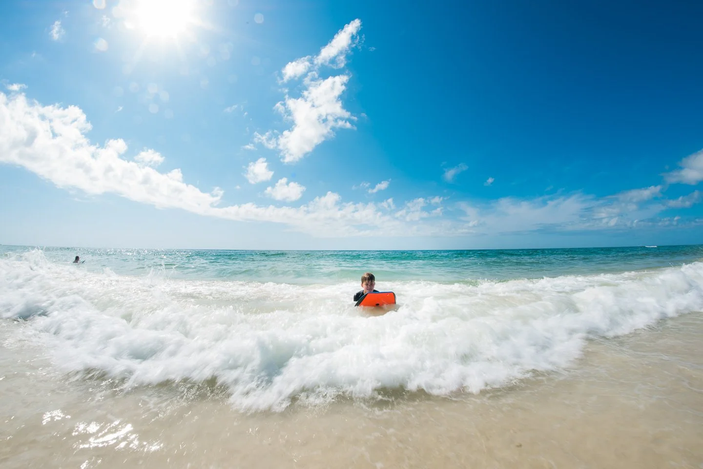 Child in the ocean with a boogie board, blue sky with clouds, sun shining, sandy beach, waves crashing.