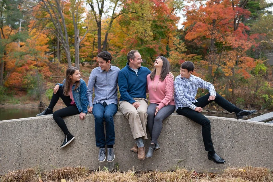 Family of five sitting on a concrete barrier outdoors during autumn, with colorful fall trees in the background.