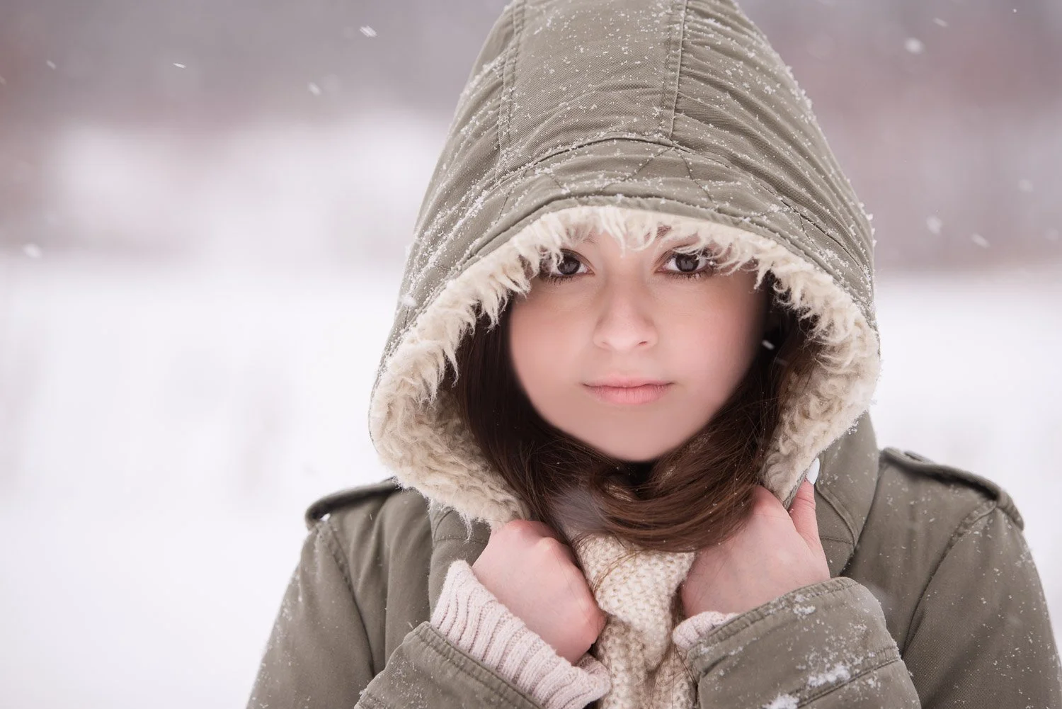 A young woman with dark hair and fair skin in a snow-covered outdoor setting, wearing a hooded winter coat with fur lining and gloves, holding the collar of her coat near her neck.