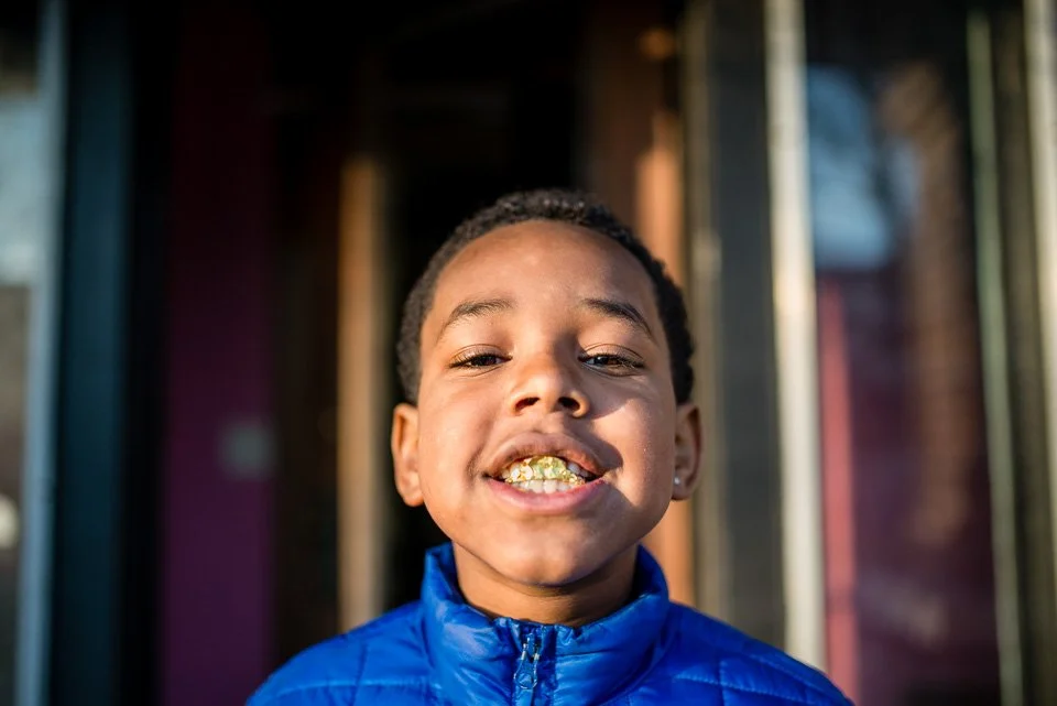 Close-up of a smiling young boy with gold teeth, wearing a blue puffer jacket, standing outside in front of a building with glass doors.