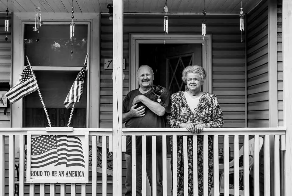 A man and woman standing on a porch with American flags and patriotic decorations, smiling at the camera.