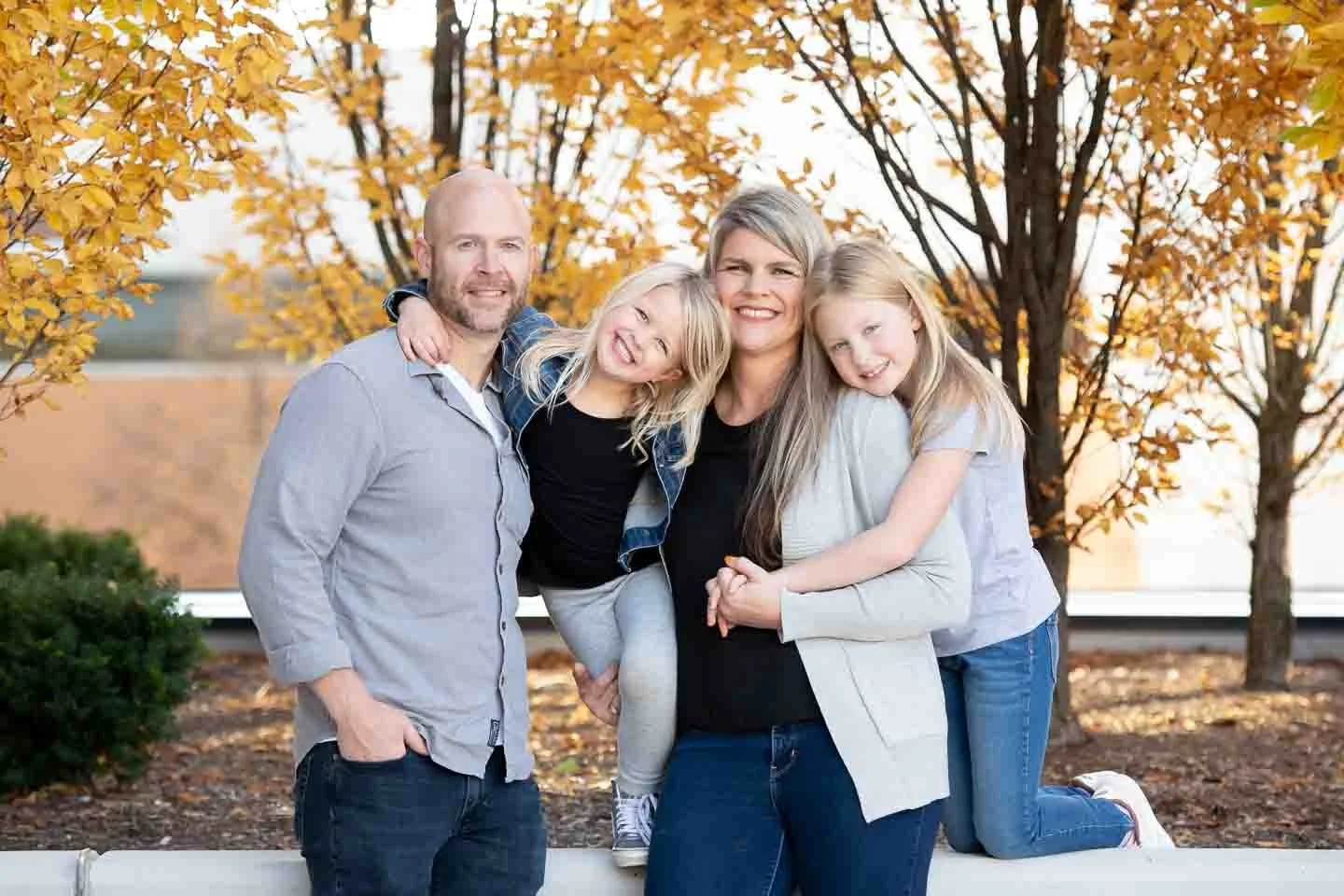 A family of four posing outdoors on a fall day in front of a tree with orange and yellow leaves. The father, mother, and two young daughters are smiling and hugging.