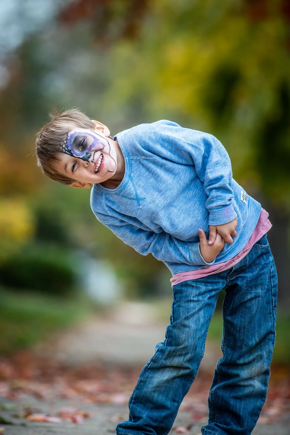 A young boy with face paint resembling a skeleton, wearing a blue sweatshirt and jeans, smiling and bending slightly while outdoors on a path surrounded by autumn foliage.