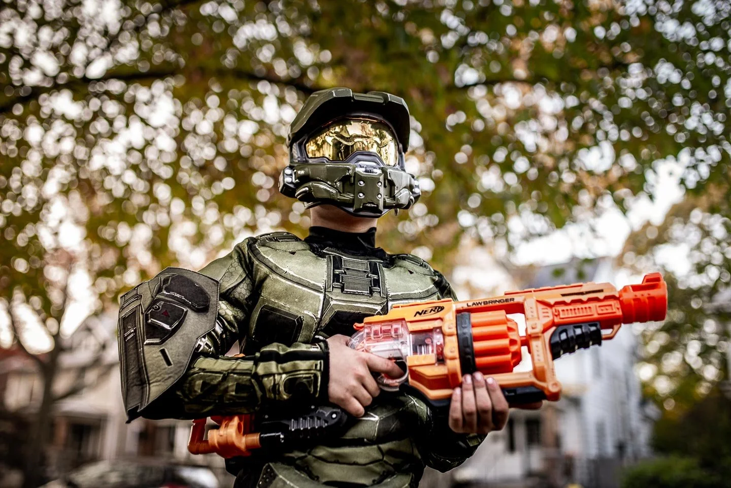 A person in a green armor costume wearing a helmet with a reflective visor, holding an orange water gun in an outdoor setting with falling autumn leaves.