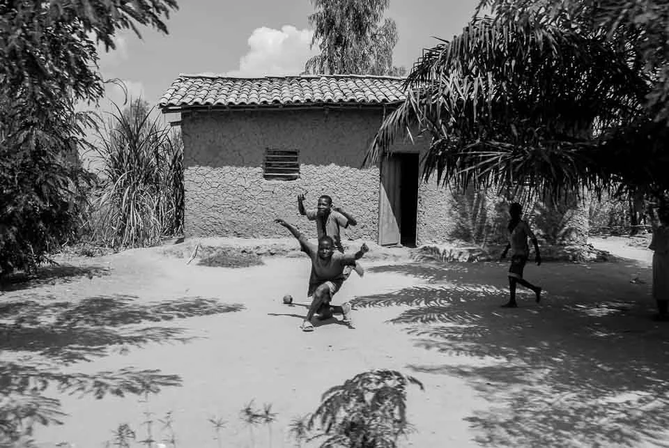 Children playing soccer outside a small house with a tile roof, surrounded by trees and shadows in a rural area.