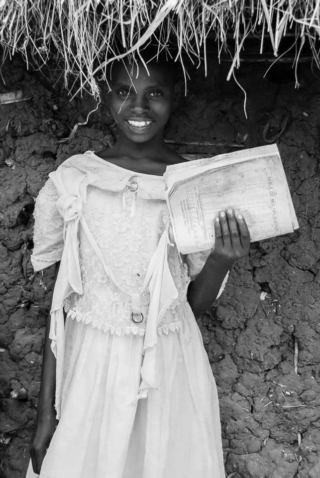 A smiling young girl stands against a dirt wall holding a book. She is wearing a light-colored dress with lace details.