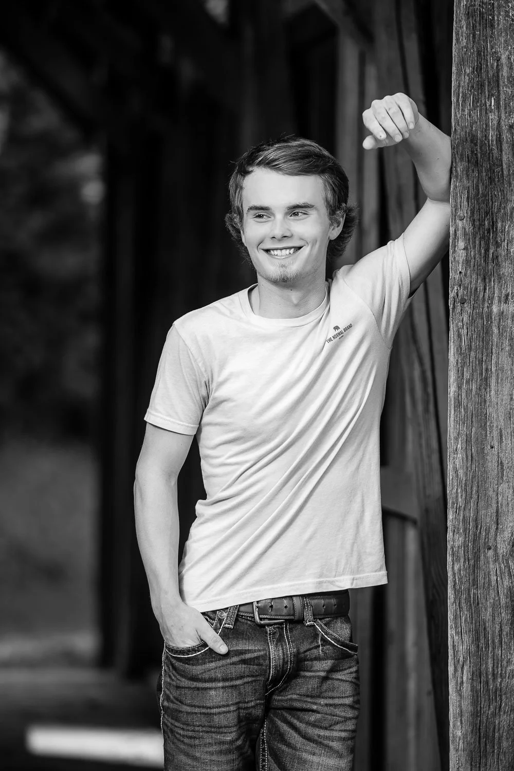 A young man with medium-length hair, smiling, wearing a light t-shirt and jeans, leaning against a wooden post outdoors.