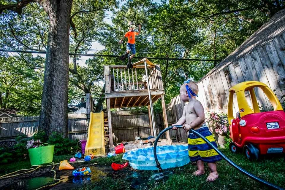 A young boy in striped swim trunks and goggles is watering a small plastic kiddie pool with a garden hose in a backyard. In the background, a toddler with a mask on walks on a wooden play structure, which has a slide. The yard is fenced, and there ar