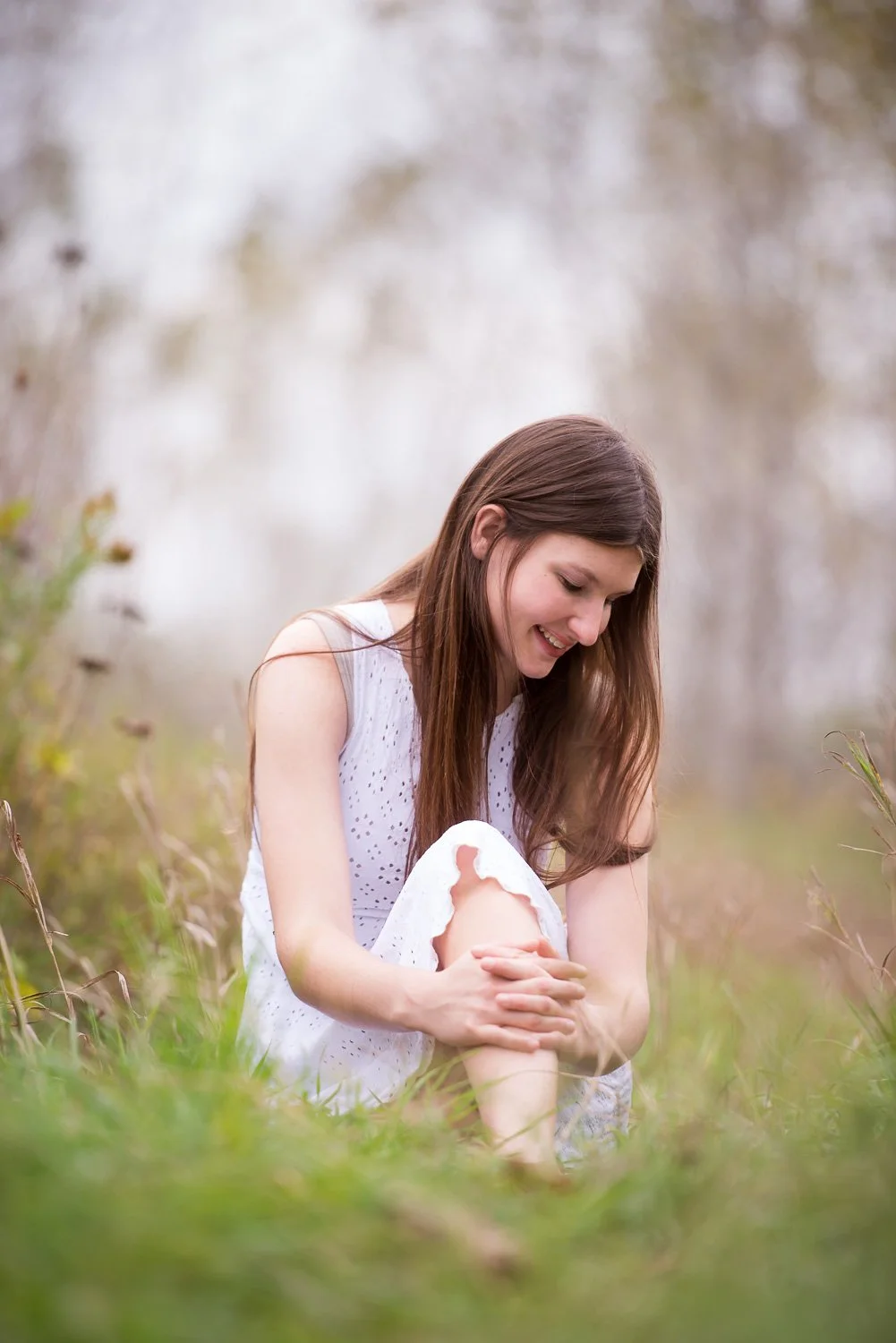 A woman sitting outdoors in a grassy area, smiling and looking down, wearing a white dress.