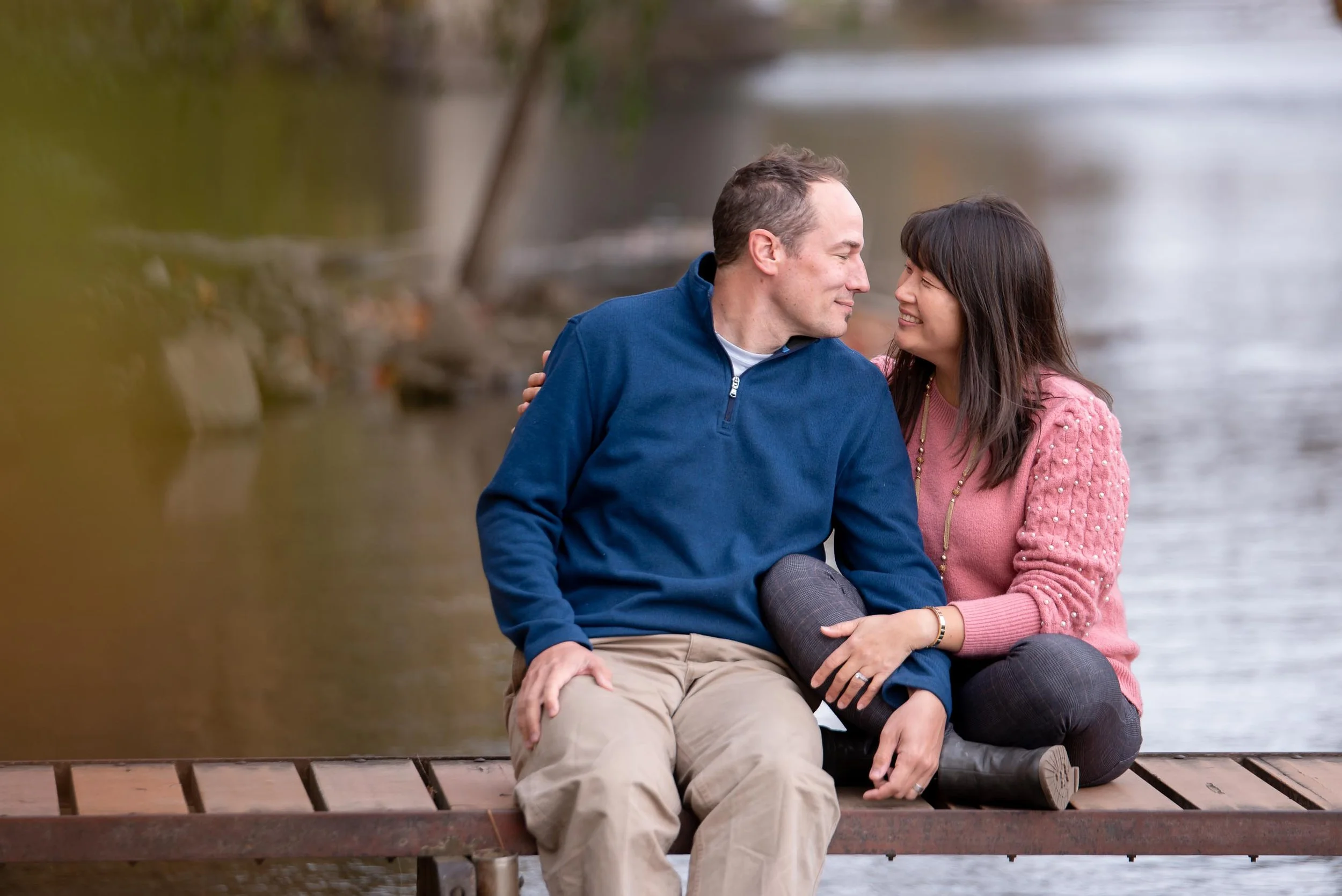 A couple sitting on a wooden dock by a river, close to each other, smiling and looking into each other's eyes.