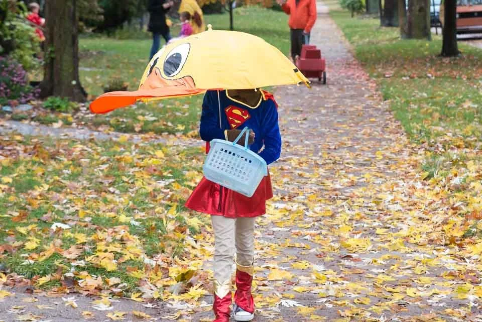 Child dressed as Supergirl holding a yellow duck-shaped umbrella and carrying a plastic basket, walking on a leaf-covered path in a park during fall.