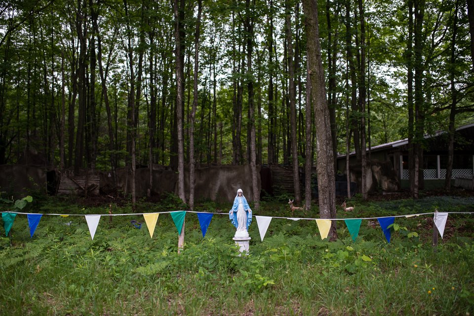 Statue of the Virgin Mary in a wooded area with colorful triangular flags strung in front and deer grazing nearby.