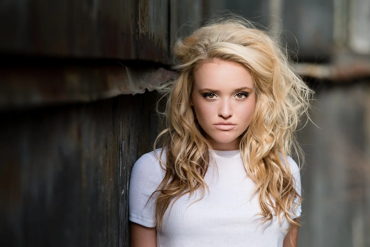 A young woman with blonde, curly hair and piercing eyes standing against a wooden wall, wearing a white shirt.