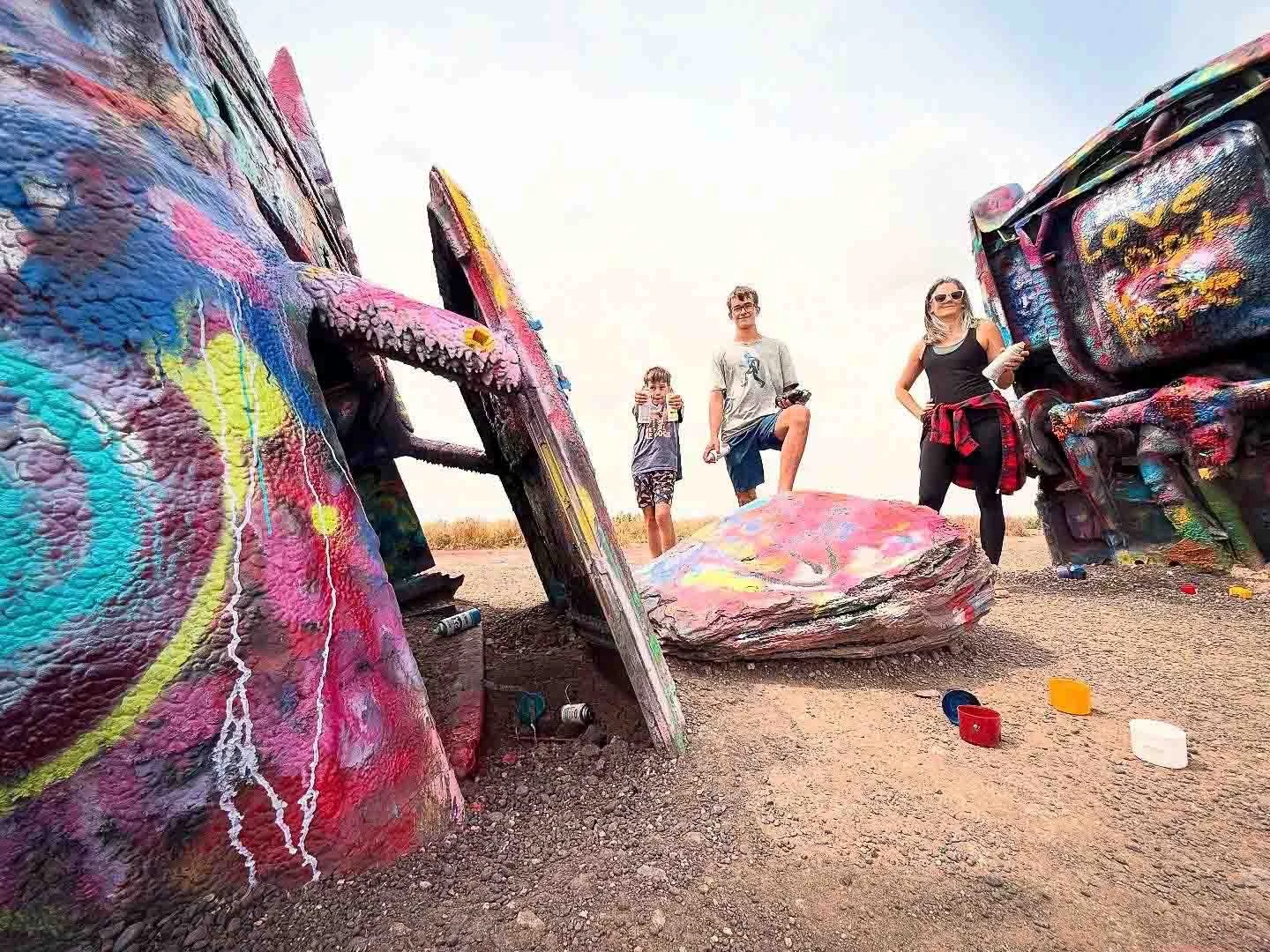 Three people, two adults and one child, stand on dirt terrain surrounded by large, colorful graffiti-covered rocks. The rocks are painted with vibrant abstract designs, with spray paint cans on the ground nearby, outdoors on a clear day.