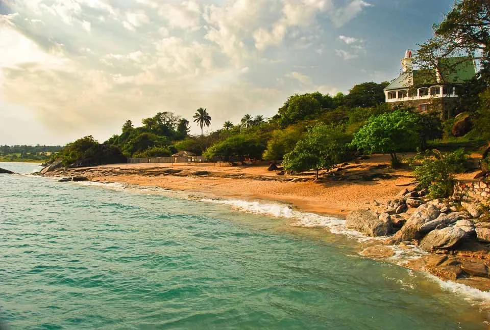 A coastal scene with a sandy beach, green trees, rocky shoreline, and a large white house on a hillside under a partly cloudy sky.