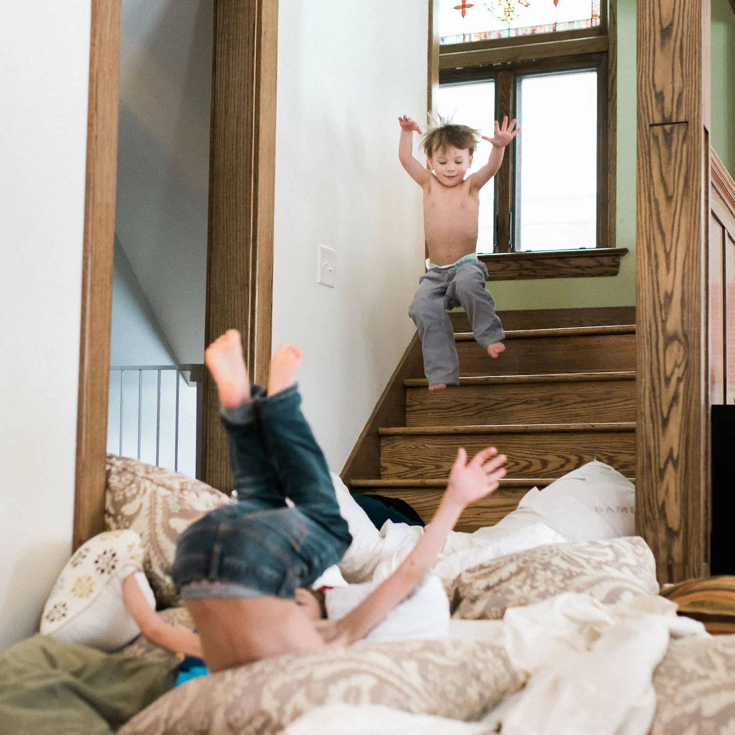 Two children playing indoors. One child is lying on a bed, upside down, with legs in the air. The other child is jumping down the stairs, with arms raised, shirtless and wearing gray pants.
