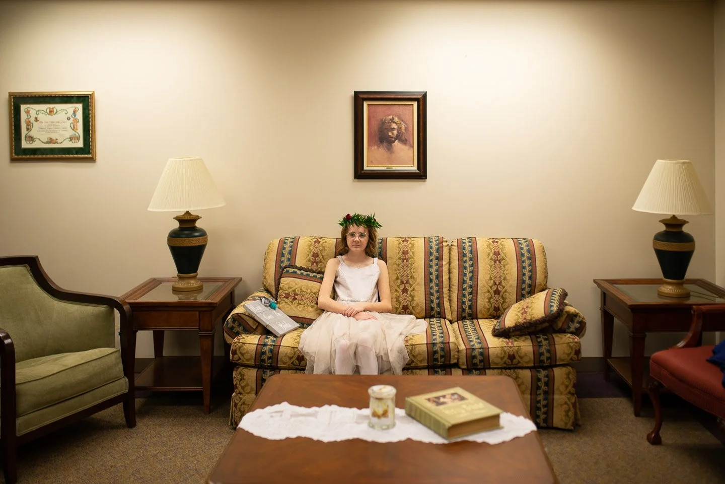 A young girl with glasses and a flower crown sitting on a patterned sofa in a living room, with lamps and framed artwork on the wall behind her.