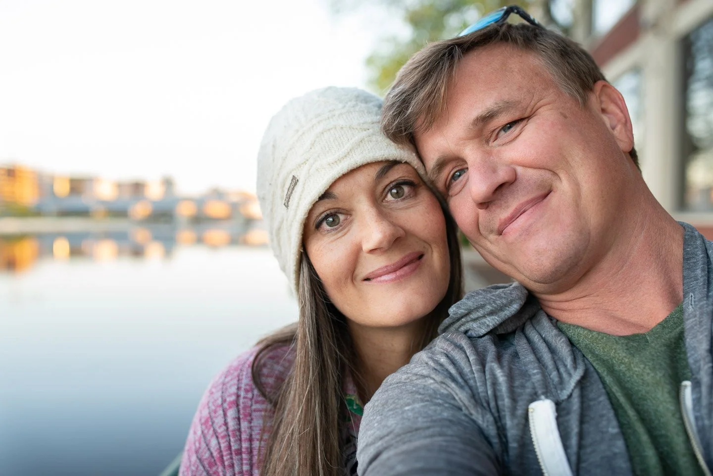 A smiling couple taking a selfie outdoors near a body of water with a cityscape in the background.