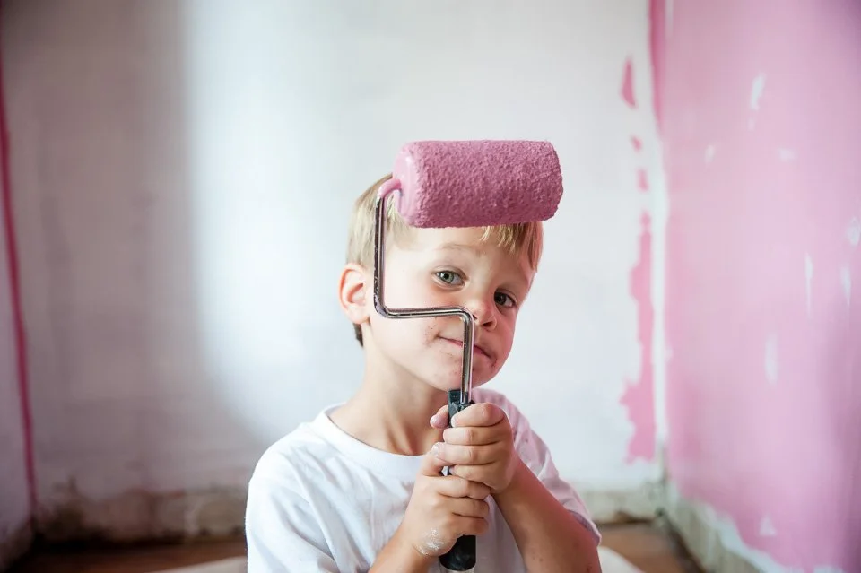 Young boy holding a paint roller with pink paint, standing in a room with partially painted pink walls.