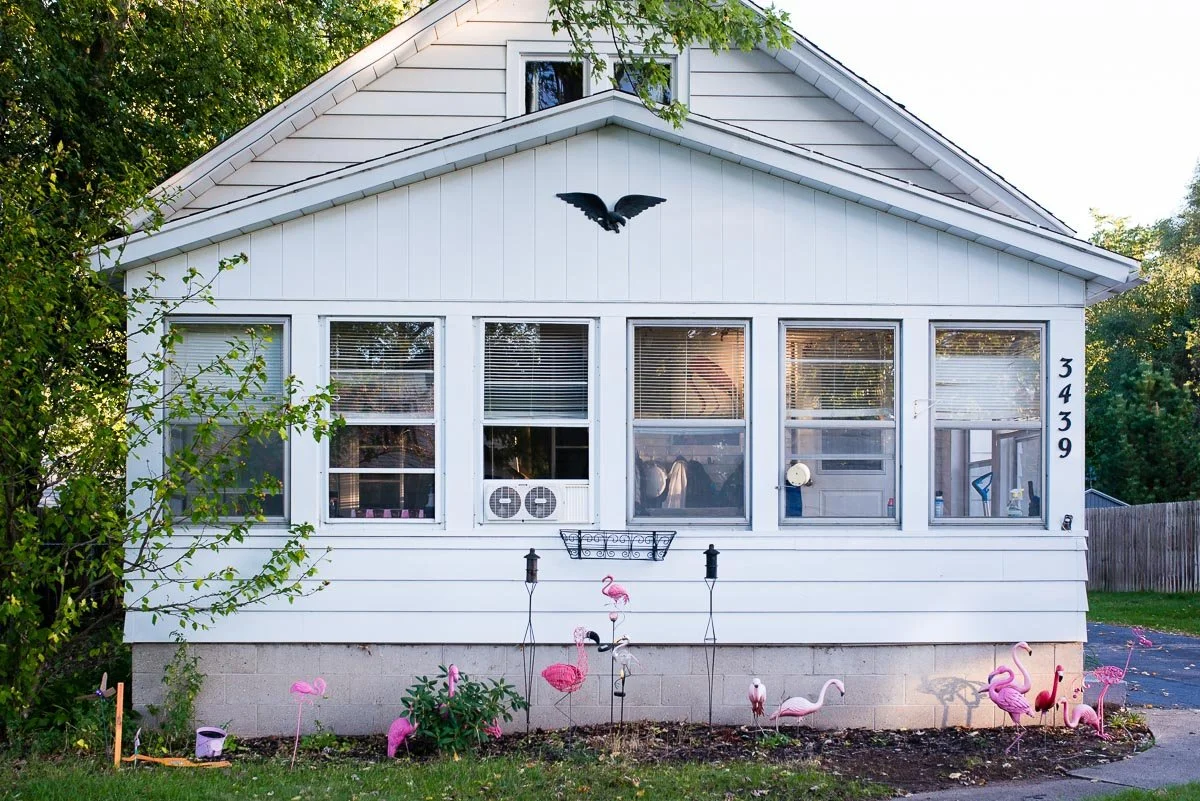 Front view of a white house with large windows, house number 3439, decorated with pink flamingo lawn ornaments, and a black bird decoration on the house's upper facade.