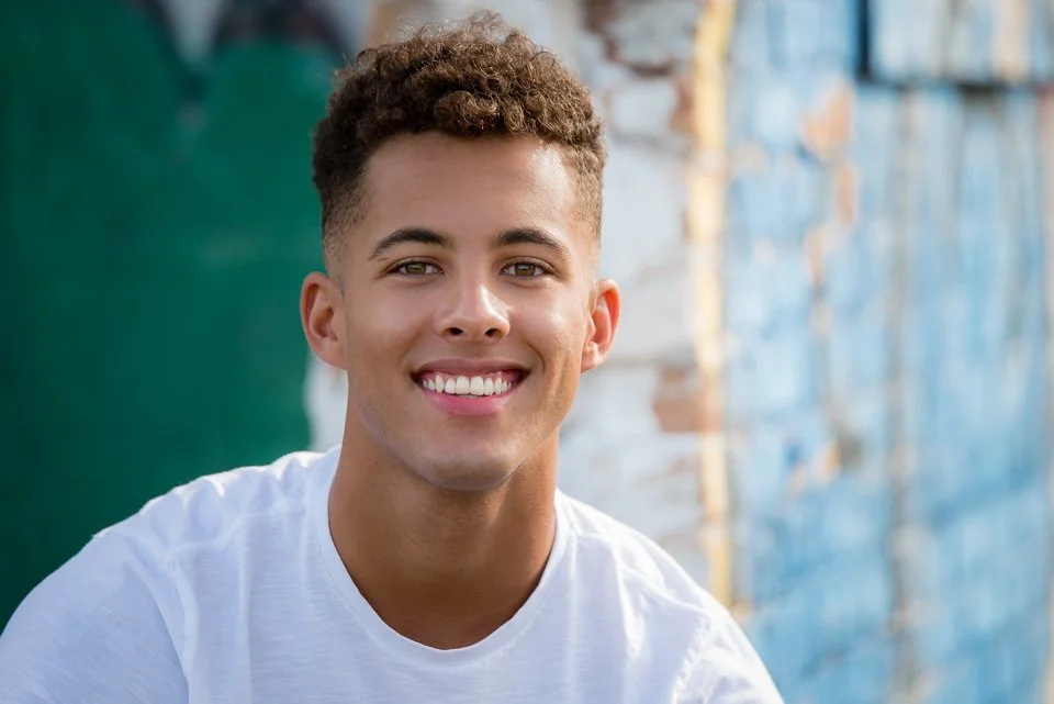 A young man with curly brown hair and a bright smile, wearing a white t-shirt, in front of a colorful outdoor background.