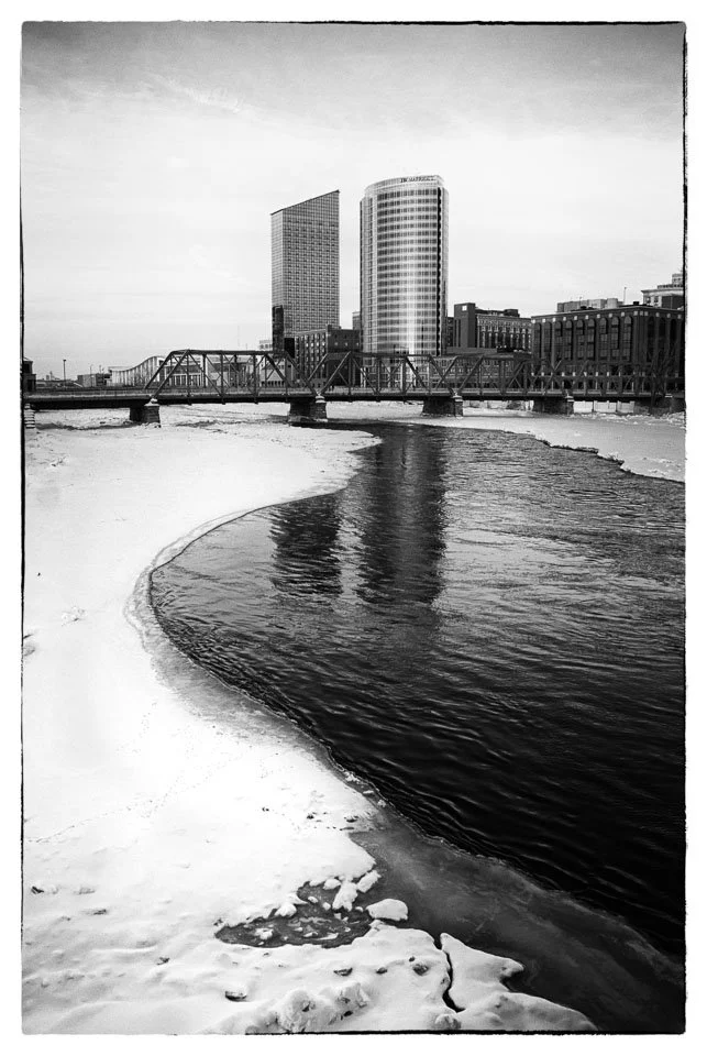 Black and white photo of a city skyline with tall buildings, a bridge, and a river with snow and ice along the banks.