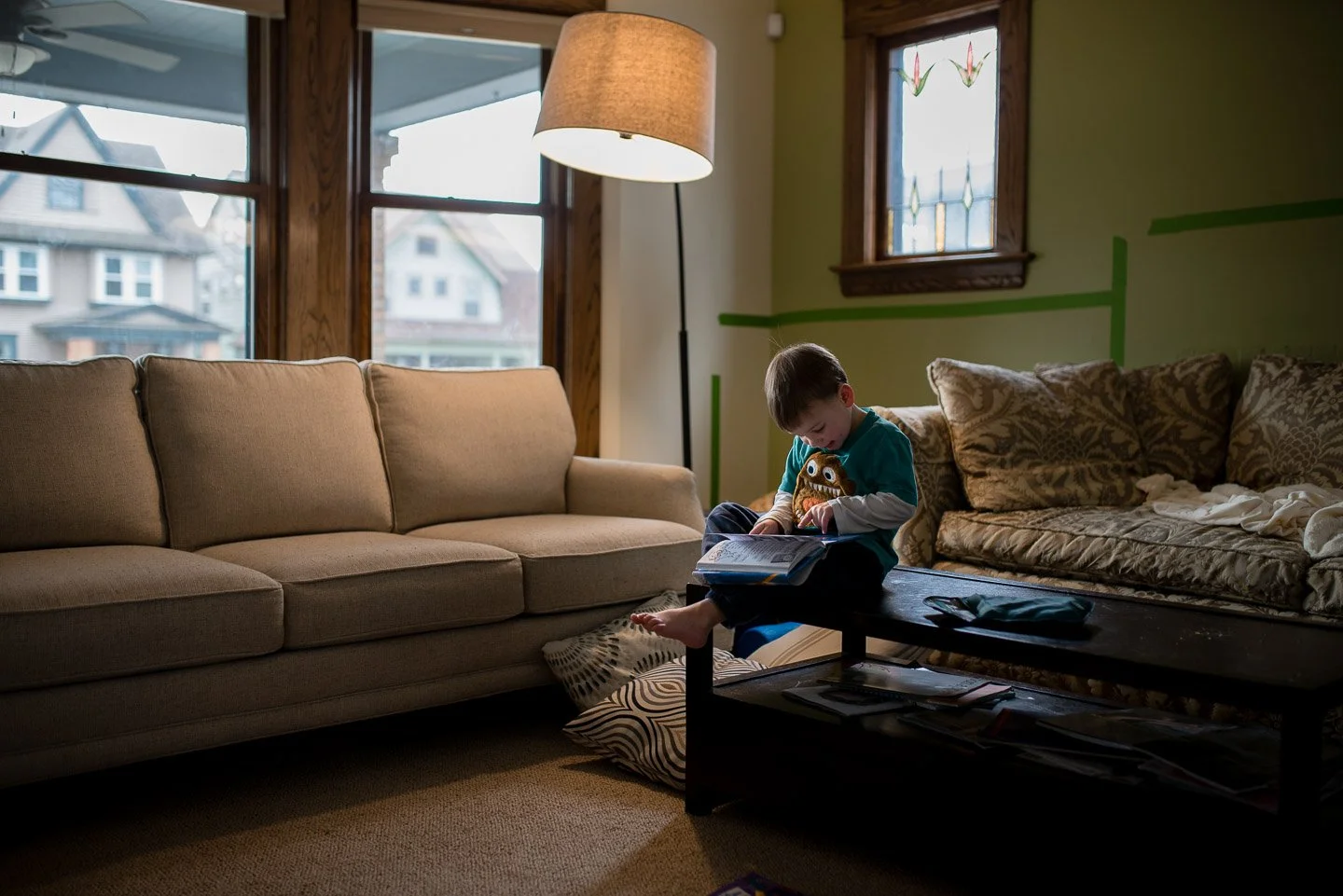 A young boy sitting on a table in a living room, reading a book and holding a plush toy. The room has large windows, a beige sofa on the left, a patterned sofa on the right, a floor lamp, and scattered books and items on the table and floor.